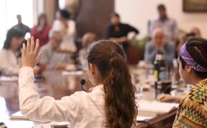 Varios niños con túnica en una mesa redonda con micrófono, levantando la mano y participando. Hay algunos adultos en segundo plano observandolos.