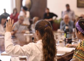 Varios niños con túnica en una mesa redonda con micrófono, levantando la mano y participando. Hay algunos adultos en segundo plano observandolos.
